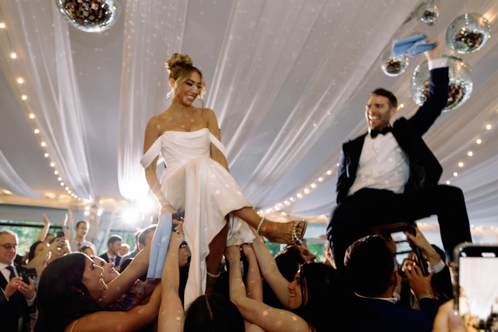 Newlyweds lifted on chairs during the hora, surrounded by dancing guests and disco balls